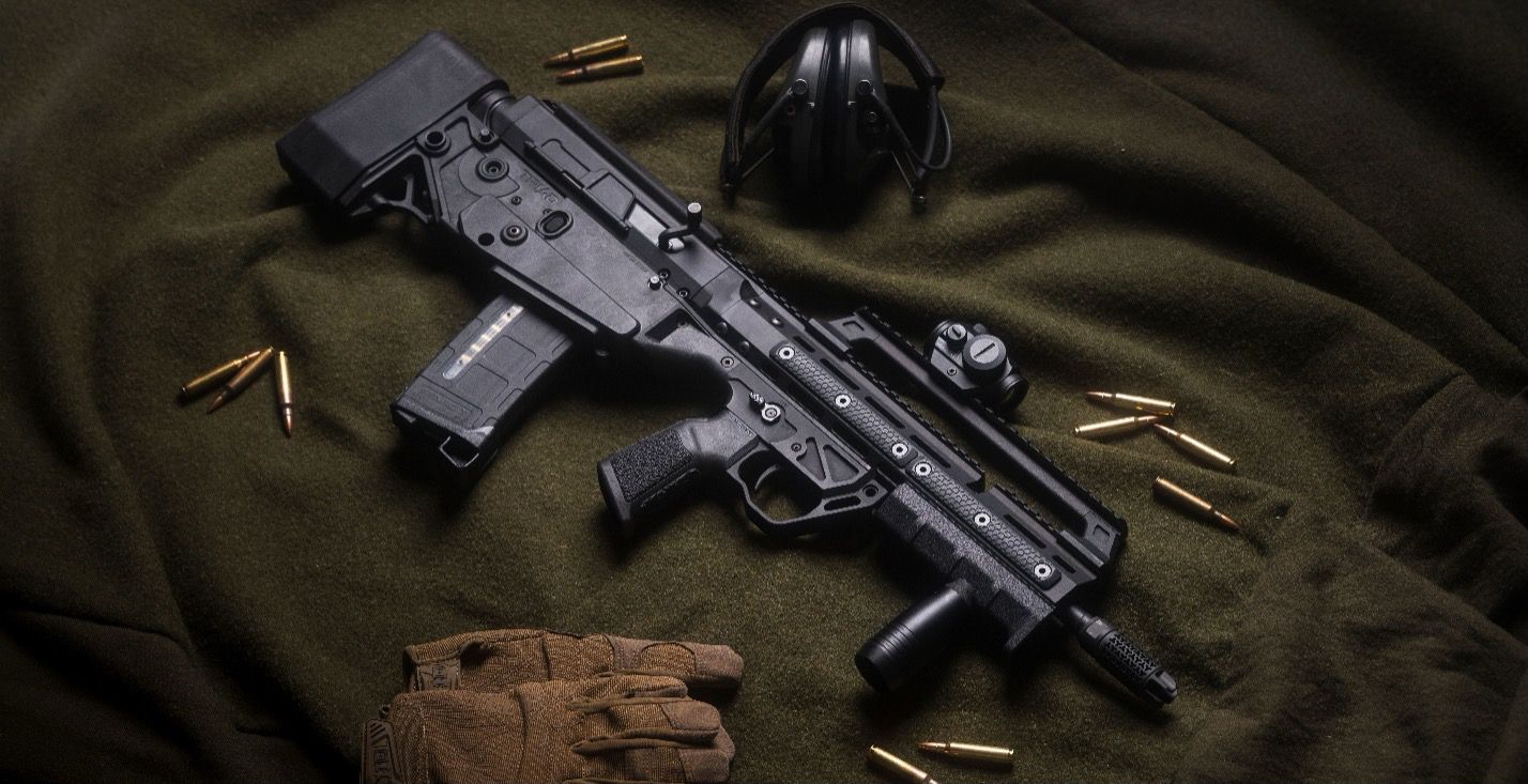 Gun with a Precision Armament EFAB&reg; resting on dark green felt surrounded by a pair of gun gloves, ear protection, and scattered bullet groups